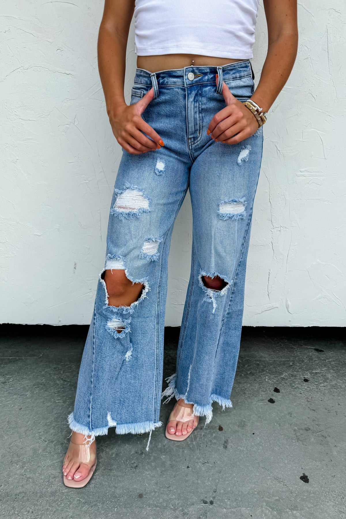Person wearing distressed blue jeans with a white top against a white wall.