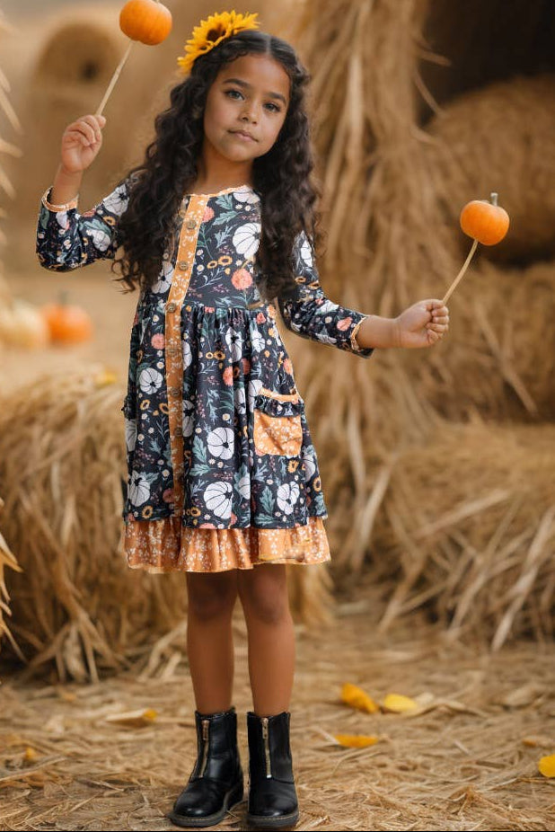 A young girl stands in a hay bale themed setting, wearing a long-sleeve floral print dress with a gold-orange front placket and hem, holding two small orange pumpkins on sticks.