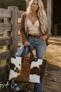 A woman stands outdoors beside a wooden fence holding a large tote bag. The bag has a cowhide-style pattern with white, brown, and black patches plus a brown tooled leather panel.