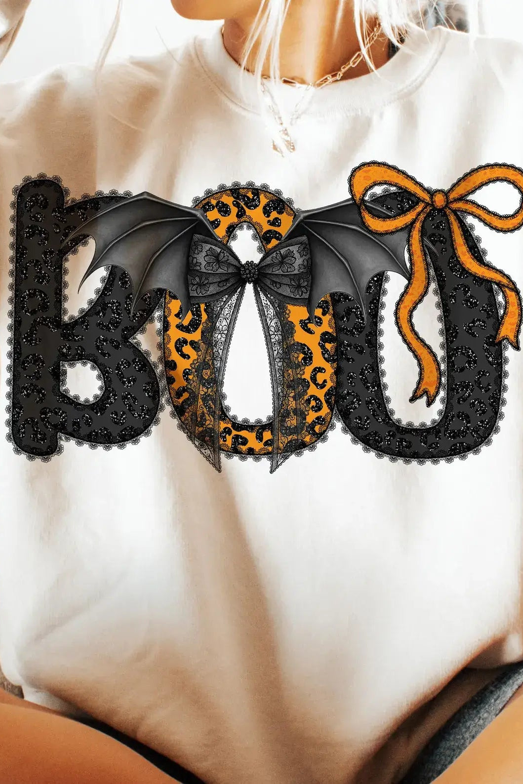 A person wearing a cream sweatshirt with a large 'BOO' graphic across the chest. The letters are black lace-textured with one in the center featuring a leopard print pattern and black bat wings, and an orange bow sits above.