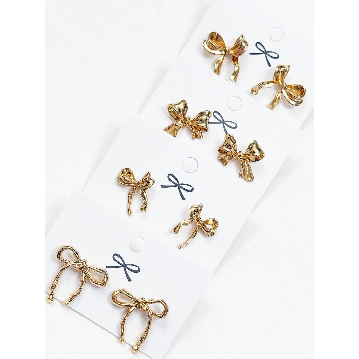 Six pairs of gold bow-shaped stud earrings displayed on a white card with printed bow illustrations, photographed on a light neutral background.