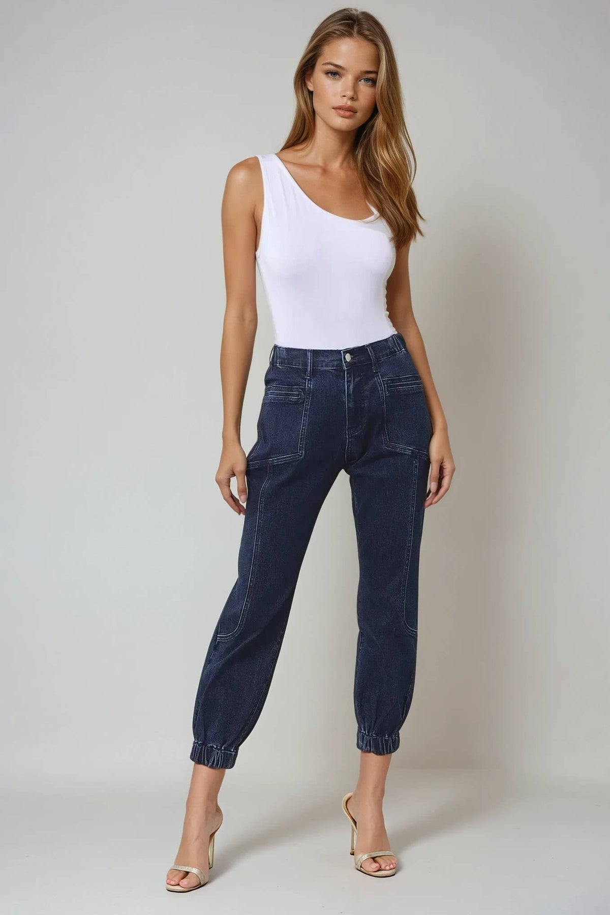 Woman wearing a white tank top and dark blue jeans against a plain background