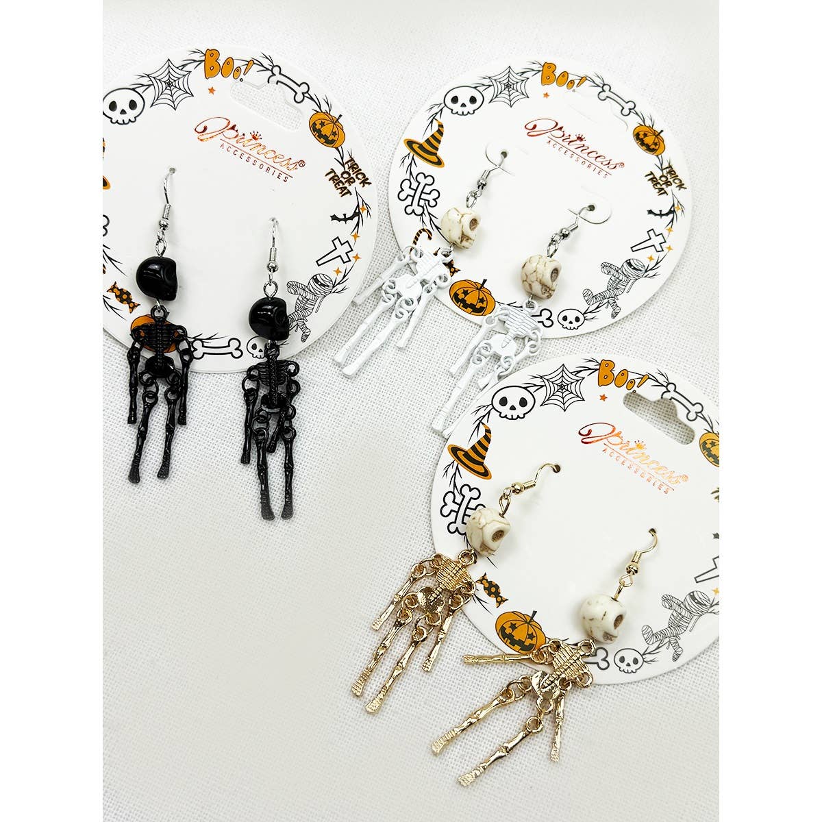 Three pairs of Halloween skeleton dangle earrings displayed on branded circular card holders. The left pair is black with orange accents, the middle pair is white with orange accents, and the right pair is metallic gold with orange accents.