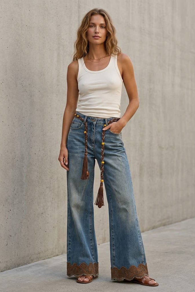 A woman stands against a concrete wall wearing wide-leg blue jeans with a brown lace hem and tassel detail, a white ribbed tank top, and brown sandals.