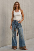 A woman stands against a concrete wall wearing wide-leg blue jeans with a brown lace hem and tassel detail, a white ribbed tank top, and brown sandals.