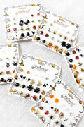 Four white display cards filled with small Halloween-themed stud earrings arranged on a light gray fabric background. The cards feature designs like witches' hats, spiders, pumpkins, bats, skulls, and ladybugs in black, orange, red, gold, and silver tones.