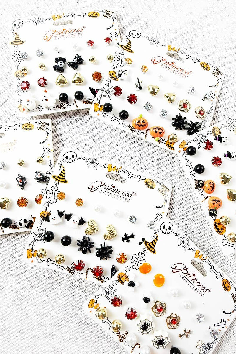 Four white display cards filled with small Halloween-themed stud earrings arranged on a light gray fabric background. The cards feature designs like witches' hats, spiders, pumpkins, bats, skulls, and ladybugs in black, orange, red, gold, and silver tones.