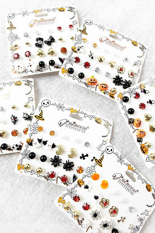 Four white display cards filled with small Halloween-themed stud earrings arranged on a light gray fabric background. The cards feature designs like witches' hats, spiders, pumpkins, bats, skulls, and ladybugs in black, orange, red, gold, and silver tones.
