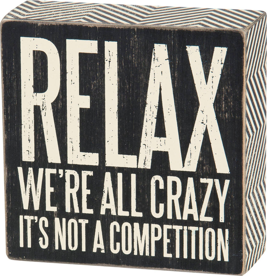 Black wooden block with white text 'RELAX WE'RE ALL CRAZY IT'S NOT A COMPETITION' on a white background