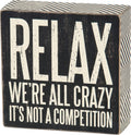 Black wooden block with white text 'RELAX WE'RE ALL CRAZY IT'S NOT A COMPETITION' on a white background