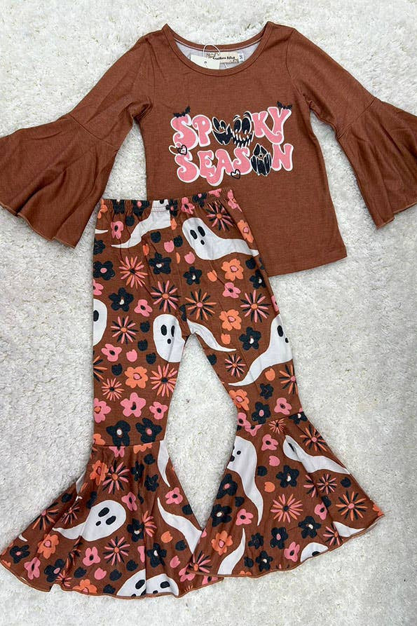A toddler girl's two-piece Halloween outfit laid flat on a white textured rug. The set includes a rust brown long-sleeve top with 'Spooky Season' printed in pink and black on the chest, and matching rust brown flared pants with a white, pink, and black ghost and floral pattern.