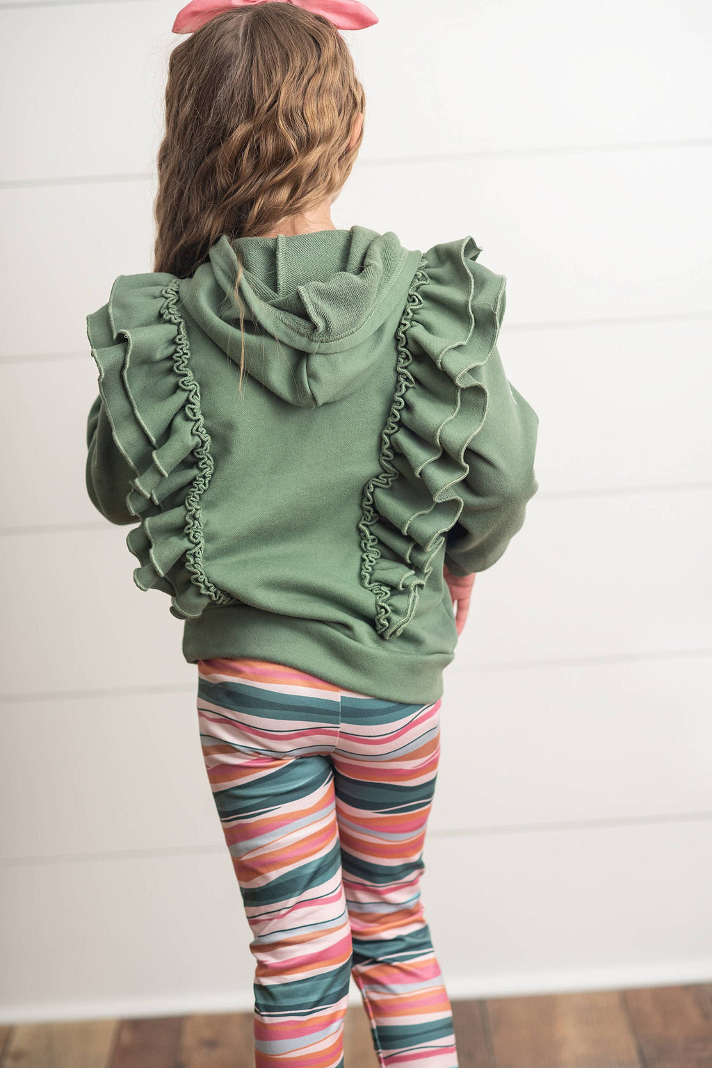 Child wearing a green hoodie with ruffles and colorful striped leggings against a white wall.