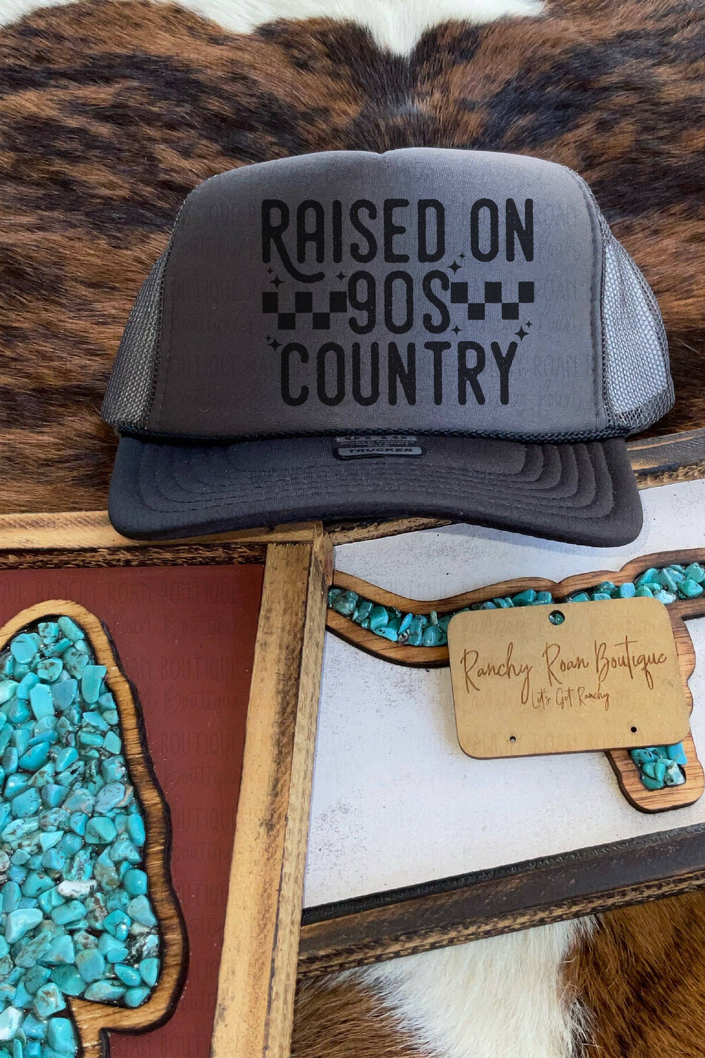 A dark gray trucker hat with 'RAISED ON 90S COUNTRY' printed on the front, displayed on a brown and white cowhide rug alongside decorative framed turquoise stone art.