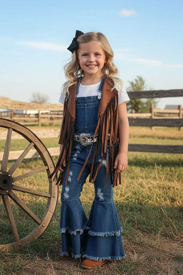 A young girl stands outdoors in a grassy field next to a large wooden wagon wheel, wearing a western-style outfit consisting of blue denim bell-bottom jeans, a brown fringed vest, a white t-shirt, and a silver concho belt.