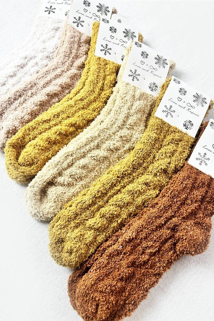 Five pairs of chunky ribbed knit socks in neutral earth tones, including cream, mustard yellow, ochre, and rust brown, arranged diagonally on a white background with decorative paper tags.