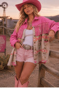 A woman posing in a rustic outdoor setting wearing a vibrant pink cowboy hat, pink fringed jacket, white tank top, light pink denim shorts, and pink cowboy boots. A wooden fence and a sign reading 'Wild Heart Ranch' are visible in the background.