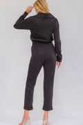 A woman shown from the back wearing a dark charcoal gray long-sleeve collared jumpsuit with an elastic waistband and cropped legs, paired with nude high-heeled sandals against a light gray studio background.