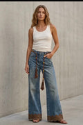 A woman stands against a concrete wall wearing a white ribbed tank top, wide-leg blue jeans with brown lace trim at the hems, and brown flat sandals.