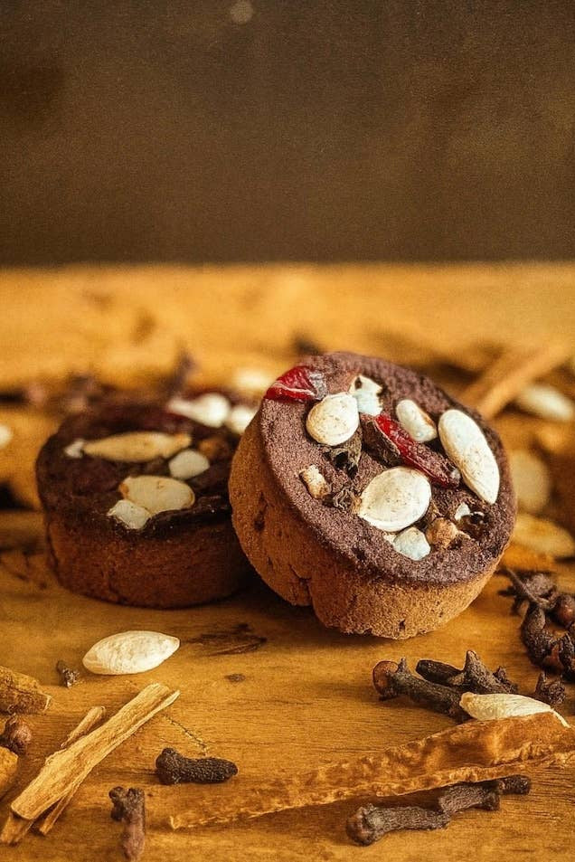 Two small round brown baked goods topped with sliced almonds, dried red chili, and spices, arranged on a wooden surface with cinnamon sticks, cloves, and seeds around them.