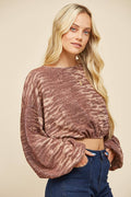 A blonde woman wearing a long-sleeve, cropped knit top with a mottled brown and beige zebra-like pattern, paired with dark blue jeans, against a solid beige background.